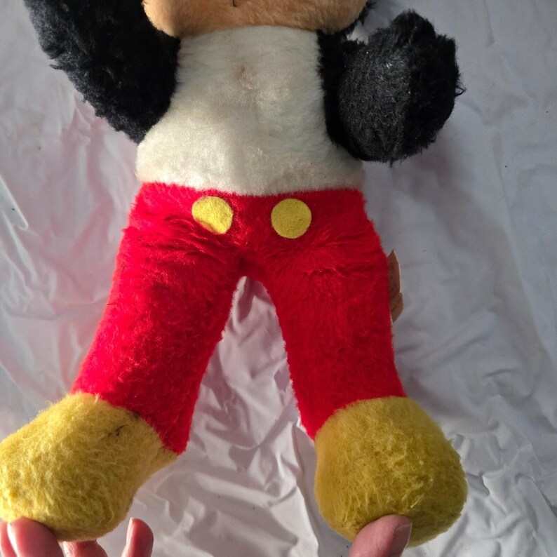 May include: Vintage Mickey Mouse plush toy. The plush features a white torso, red pants with yellow buttons, black arms, and yellow feet. The toy's arms are raised, and it appears to be in good condition for its age.