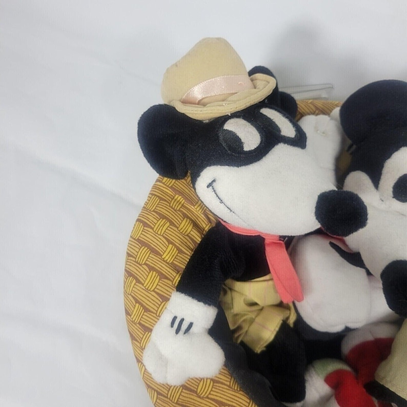 May include: A collection of Mickey Mouse plush toys. One Mickey Mouse is wearing a tan hat with a pink ribbon, a red scarf, and a yellow vest. The plush toys are in a basket with a woven pattern.