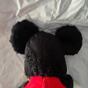 May include: A plush toy featuring a black head with large, round ears and a red torso. The toy's black fur is textured, and the ears are prominently displayed against a white background. The toy is a classic character.