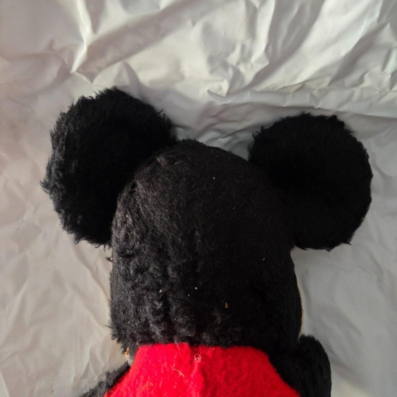 May include: A plush toy featuring a black head with large, round ears and a red torso. The toy's black fur is textured, and the ears are prominently displayed against a white background. The toy is a classic character.