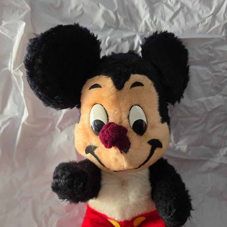 May include: A vintage Mickey Mouse plush toy. The plush features a tan face, black ears, and a red nose. Mickey is wearing red shorts with yellow buttons. The toy has black arms and a white belly.