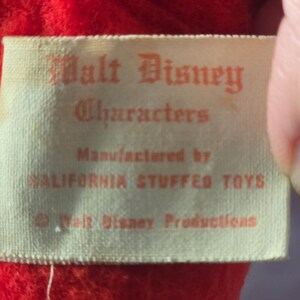 May include: Close-up of a vintage tag with the text "Walt Disney Characters" in red lettering. The tag is cream-colored and reads "Manufactured by CALIFORNIA STUFFED TOYS" and "&copy; Walt Disney Productions."