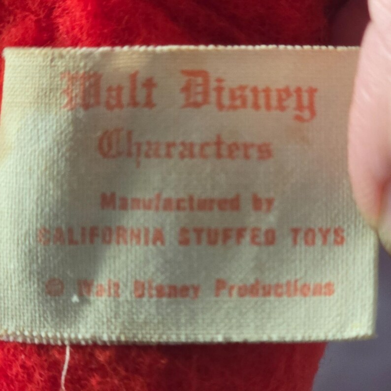 May include: Close-up of a vintage tag with the text "Walt Disney Characters" in red lettering. The tag is cream-colored and reads "Manufactured by CALIFORNIA STUFFED TOYS" and "&copy; Walt Disney Productions."