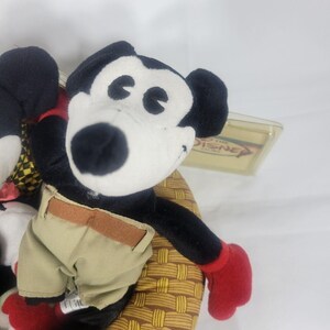 May include: A plush Mickey Mouse toy with black fur, a white face, and red gloves. Mickey wears khaki shorts with a brown belt. The toy is seated on a woven basket. A Disney tag is visible.
