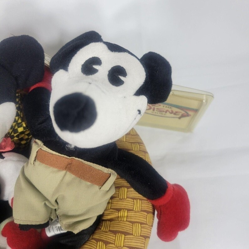 May include: A plush Mickey Mouse toy with black fur, a white face, and red gloves. Mickey wears khaki shorts with a brown belt. The toy is seated on a woven basket. A Disney tag is visible.