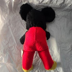 May include: Vintage Mickey Mouse plush toy, viewed from the back. The plush features black fur for the head and ears, red pants, and yellow feet. The toy is on a white, textured surface.