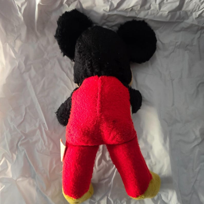 May include: Vintage Mickey Mouse plush toy, viewed from the back. The plush features black fur for the head and ears, red pants, and yellow feet. The toy is on a white, textured surface.