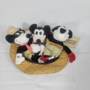 May include: Three Mickey Mouse plush toys in a woven basket. The Mickey Mouse toys are black, white, and tan, with red and tan clothing. One Mickey Mouse toy wears a cowboy hat. A Disney tag is visible.