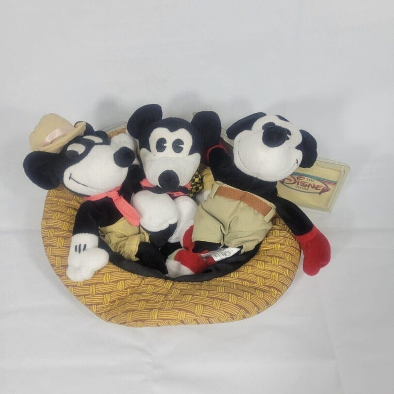 May include: Three Mickey Mouse plush toys in a woven basket. The Mickey Mouse toys are black, white, and tan, with red and tan clothing. One Mickey Mouse toy wears a cowboy hat. A Disney tag is visible.