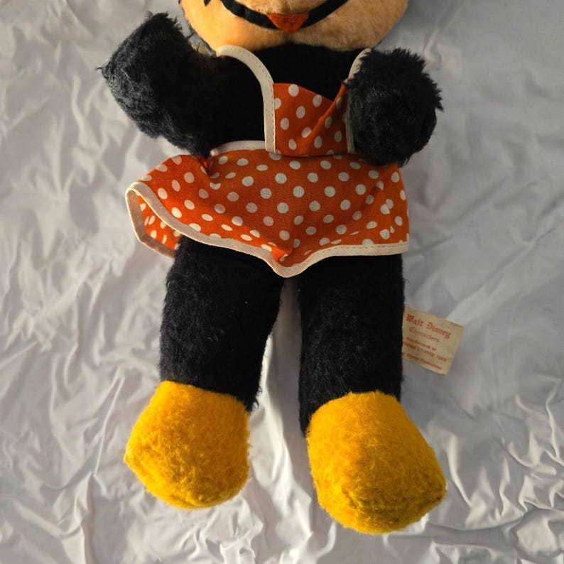 May include: Vintage Minnie Mouse plush doll with black fur, orange and white polka dot dress, yellow feet, and a tag that reads "Walt Disney Character". The doll has black arms and legs and a smiling face.