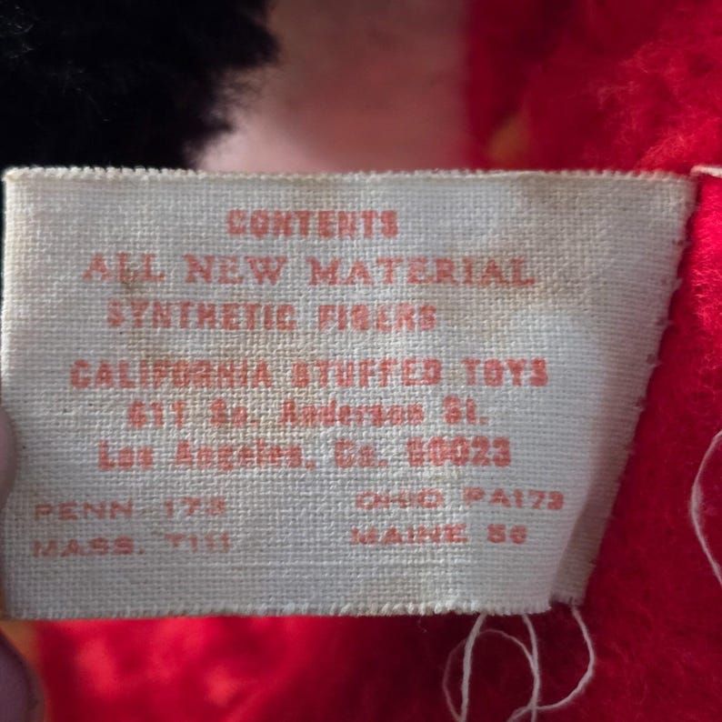 May include: Close-up of a rectangular tag with red text on a cream background. The tag reads "CONTENTS ALL NEW MATERIAL SYNTHETIC FIBERS CALIFORNIA STUFFED TOYS". Additional text includes an address and state abbreviations.