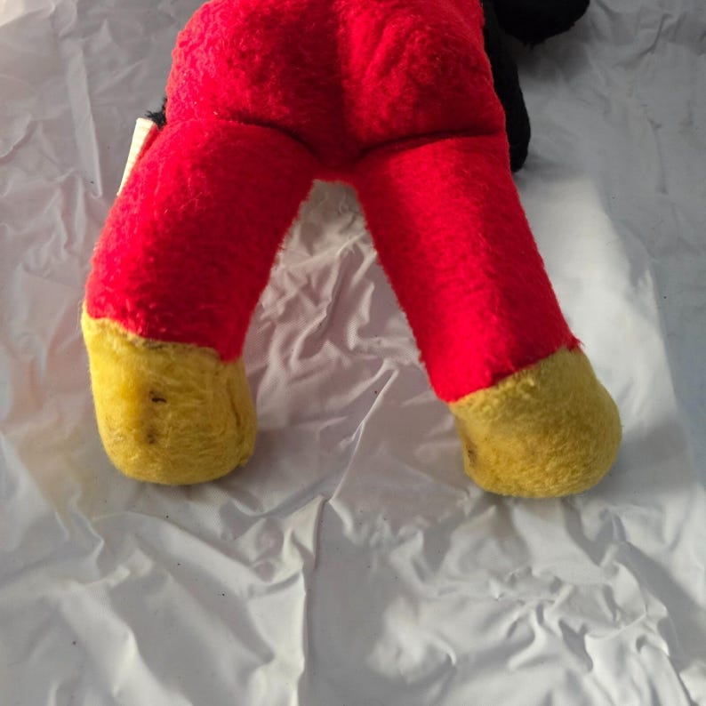May include: A vintage plush toy featuring red legs with yellow feet. The toy's legs are bent, suggesting a seated or playful pose. The plush material appears worn, indicating its age.