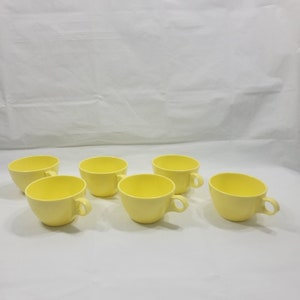 May include: Six yellow ceramic teacups with handles. The cups are arranged in a row on a white surface.