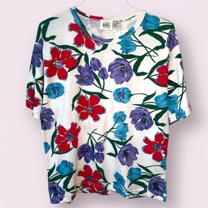 Puede incluir: Camiseta blanca de manga corta con un vibrante estampado floral. El diseño presenta grandes flores rojas, azules y moradas con tallos y hojas verdes. La camiseta está colgada de una percha blanca.
