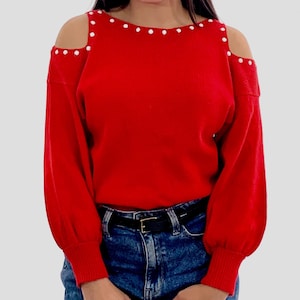 May include: A bright red sweater with open shoulders and long sleeves. The neckline and shoulder openings are adorned with small, white, circular embellishments. The sweater is paired with blue jeans and a black belt.