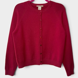 May include: A long-sleeved, magenta-colored knit cardigan featuring a round neckline and a front button-down closure with matching magenta buttons. The garment has ribbed cuffs and a ribbed hem, with a Gap brand label visible at the inner neck.