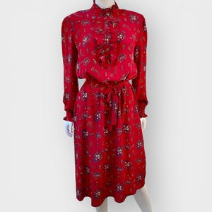 May include: A red dress with a paisley pattern. The dress has long sleeves, a high collar, and a tie waist. The dress is knee-length and has a ruffled detail on the bodice.