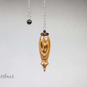 May include: A wooden pendulum with a brown and yellow wood grain pattern, hanging from a silver chain with a black bead at the top. The pendulum is shaped like a teardrop with a pointed tip. The background is a light gray.