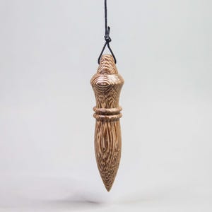 Peut inclure: Un pendule en bois brun et beige avec un motif de grain distinct, présentant une forme effilée et pointue avec deux anneaux horizontaux autour du centre, suspendu à un fin cordon noir noué en boucle au sommet.