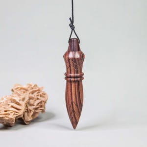 May include: A dark brown wooden pendulum with a pointed tip and carved leaf-like patterns, suspended by a thin black cord. Beside the pendulum sits a light tan, textured desert rose crystal formation with layered, petal-like structures.