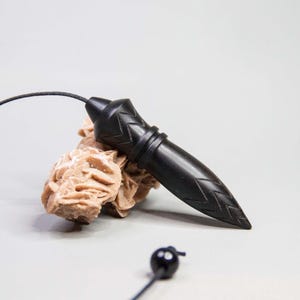 Puede incluir: Un péndulo de madera negra tallada, puntiagudo y con un patrón de chevrón, unido a un cordón negro fino. El péndulo descansa sobre una formación de cristal de rosa del desierto de color tostado claro, con una pequeña cuenta negra en el cordón.