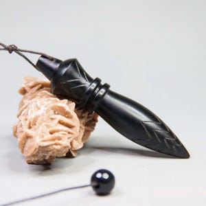 Può includere: Un ciondolo in legno nero intagliato con un'estremità appuntita e incisioni decorative, sospeso da un cordino. Poggia su una formazione di pietra strutturata marrone chiaro, con una piccola perla nera alla fine del cordino.
