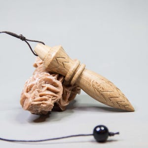 Op de afbeelding: Een lichtgekleurde houten pendel met gesneden details, verbonden met een zwart touwtje en een zwarte kraal. De pendel rust op een cluster van lichtbruine, getextureerde minerale formaties.