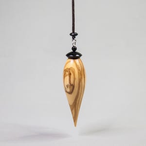 Puede incluir: Un péndulo de madera con forma de lágrima, con vetas de color marrón claro y oscuro. El péndulo está suspendido por un cordón oscuro y una pequeña cuenta negra. La parte superior del péndulo tiene una tapa negra.