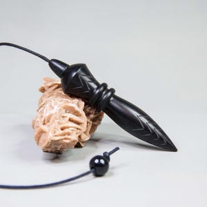 May include: A black pendulum with a pointed end and decorative carvings, attached to a black cord and a small black bead. The pendulum rests on a light brown, textured stone formation.