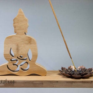 Puede incluir: Porta incienso de madera con una figura de Buda tallada con un símbolo Om y un quemador de incienso de loto de cobre. El porta incienso está hecho de bambú natural y tiene el texto "lesartsbois" grabado en la base.