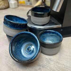Könnte beinhalten: Eine Sammlung handgefertigter Keramikbecher. Jeder Becher hat ein zweifarbiges Design mit einem grauen Sockel und einem glänzenden, tiefblauen Innenraum und Rand. Die Becher sind gestapelt und angeordnet.