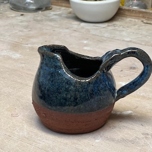 Può includere: Una piccola caraffa in ceramica fatta a mano con una glassa blu scuro su una base marrone rossiccio. La caraffa ha un corpo arrotondato, un manico e un beccuccio. Probabilmente viene utilizzata per servire panna o latte.