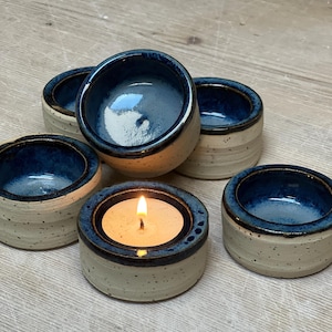 Può includere: Un set di cinque piccoli portacandele rotondi in ceramica con esterno color crema maculato e interno smaltato in blu profondo lucido. Un portacandele contiene una candelina accesa, mentre gli altri sono vuoti.