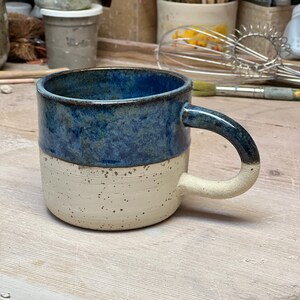 Peut inclure: Une tasse en céramique faite à la main avec une base blanc cassé mouchetée et une partie supérieure bleue texturée. La tasse a une anse incurvée et un intérieur bleu foncé.