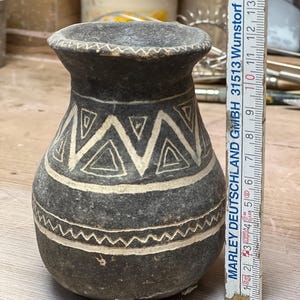 Könnte beinhalten: Eine dunkelgraue Keramikvase mit weißen geometrischen Mustern. Die Vase hat einen breiten Boden, einen schmalen Hals und einen ausgestellten Rand. Die weißen Designs umfassen Dreiecke und Zickzacklinien. Die Vase ist etwa 10 cm hoch.
