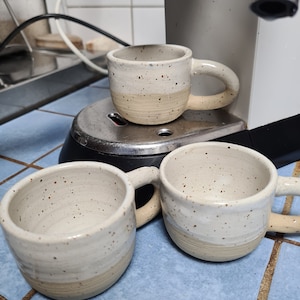 Könnte beinhalten: Drei handgefertigte Keramikbecher mit einer gesprenkelten, cremefarbenen Glasur und einem beigefarbenen Boden. Ein Becher steht auf einem silbernen Gerät, während die anderen beiden auf einer blau gefliesten Oberfläche stehen. Jeder Becher hat einen abgerundeten Henkel.