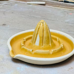 Könnte beinhalten: Handgefertigte Zitruspresse aus Keramik mit gelber Glasur. Sie hat einen konischen Reiber und eine flache Schale mit Ausguss. Das Design ist einfach und funktional, ideal für die Herstellung von frischem Saft.