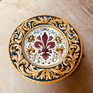 Può includere: Piatto decorativo in ceramica con un motivo a giglio al centro. Il piatto presenta uno sfondo bianco con un giglio di colore marrone, circondato da un bordo blu e oro ornato. Il bordo del piatto è giallo, circa 25 cm di diametro.