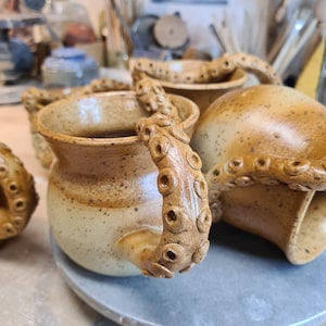 Può includere: Tre tazze in ceramica con smalto macchiato marrone e beige. Ogni tazza ha un'unica maniglia a forma di tentacolo di polpo.