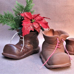 Könnte beinhalten: Drei braune Keramik-Stiefel mit Schnürsenkeln, die als Pflanzgefäße verwendet werden. Der größte Stiefel hat eine rote Weihnachtsstern-Pflanze darin.