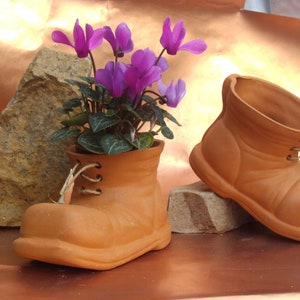 Könnte beinhalten: Zwei terracotta-Blumentöpfe in Form von Stiefeln. Der Stiefel links hat eine violette Blütenpflanze drin. Der Stiefel rechts ist leer.