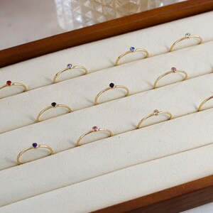 May include: A wooden box with a white velvet interior displays a collection of gold rings with colorful gemstones. Each ring features a single gemstone in a variety of colors, including red, blue, green, pink, and black.