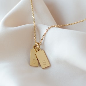 May include: A gold chain necklace with two small rectangular pendants, each engraved with a letter, "D" and "S". The necklace is displayed on a white fabric background.