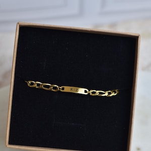 May include: A gold chain bracelet with a rectangular nameplate inside a brown box with a black interior. The nameplate has the initials "R | Z" engraved on it. The bracelet is made of gold-colored metal.