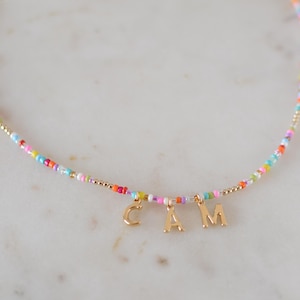 May include: A colorful beaded necklace with a gold charm that spells out the name "CAM".