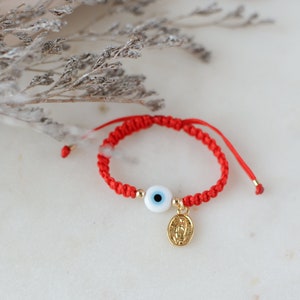 May include: Red string bracelet with a blue evil eye charm and a gold charm with a religious image.