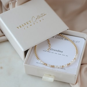 May include: A white box with a gold inscription that reads "VELVET Collection JEWELRY". Inside the box is a gold chain bracelet with small gold beads and white pearls. The bracelet is displayed on a white card with the text "Morse Code Bracelet" and "Grandma" in gold lettering. The card also includes the text "14k Gold & Pearls" and a website address.