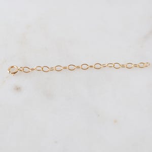 May include: A gold-colored chain extender with an oval link design. The extender has a lobster clasp on one end and a series of oval links on the other. The chain is displayed on a white marble surface.