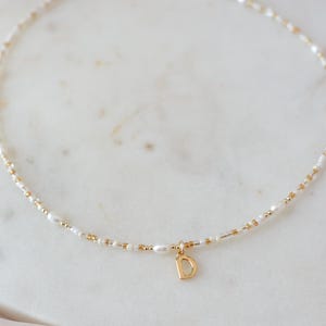 May include: Delicate necklace featuring a strand of small white pearls and gold-colored beads. A gold-colored pendant with the letter "D" hangs from the center. The necklace is displayed on a white marble surface.