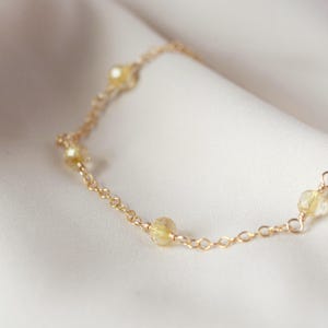 May include: A delicate gold chain bracelet with small, round, yellow gemstones evenly spaced along its length. The bracelet is displayed on a soft, white fabric, creating a subtle contrast.